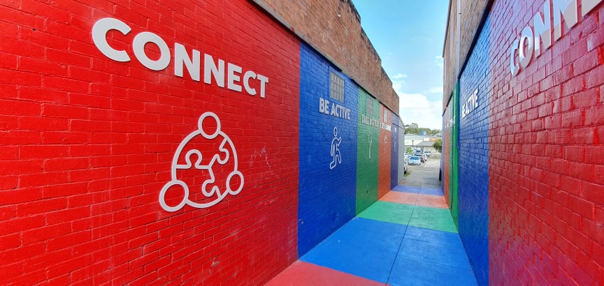 The 5 Ways to Wellbeing Laneway in Cessnock, with Connect and Be Active painted on red, blue and green brick walls above a brightly painted path