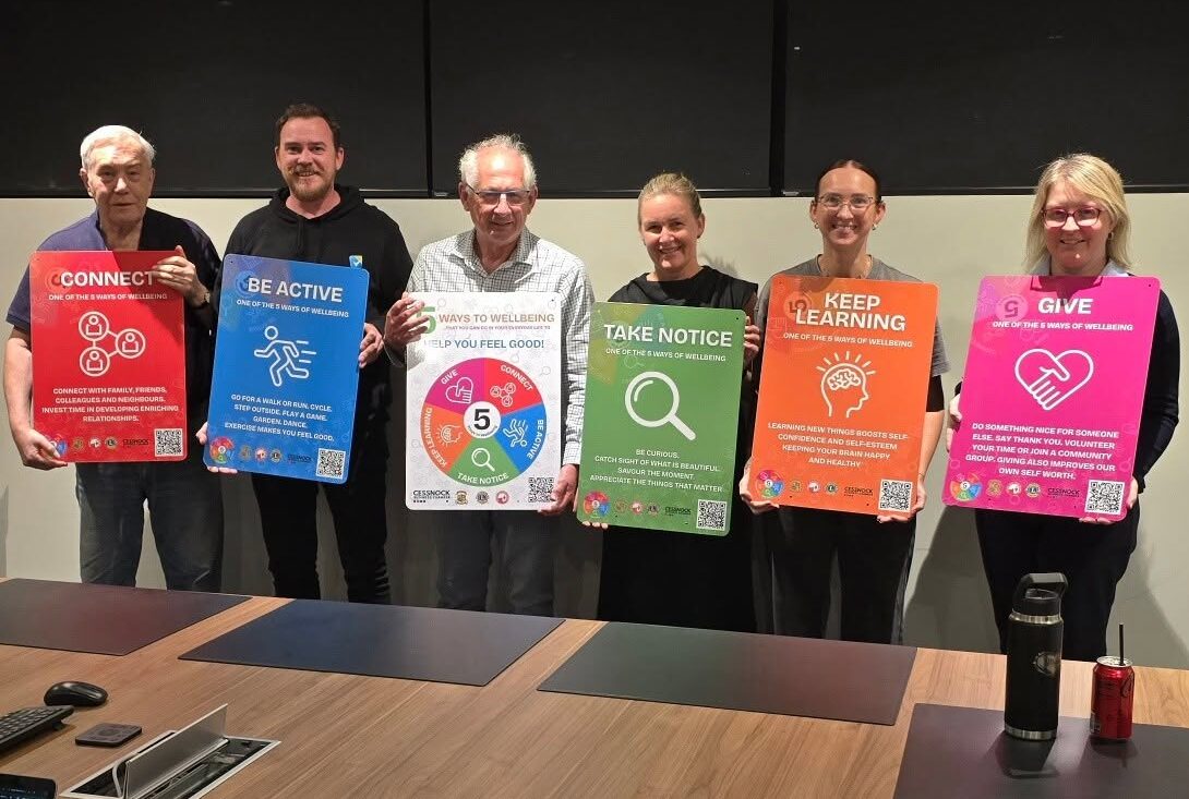 Six community members holding the 5 Ways to Wellbeing signs — Connect, Be Active, Take Notice, Keep Learning and Give